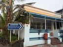 A white building with blue trim stands behind a low wall topped with metal bars, with a blue and white directional sign on posts beside a palm tree, a red post box mounted on the wall, and steps leading up to a white gate.