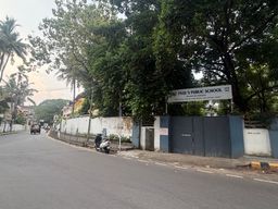 A paved road curves gently to the left, bordered by a white wall topped with a metal fence and dense green trees. A large white sign mounted above a pair of closed metal gates identifies a school, with smaller text beneath it. A scooter is parked on the sidewalk near the gate, and a vehicle drives away in the distance under an overcast sky. Palm trees rise above the foliage on the left side of the road.