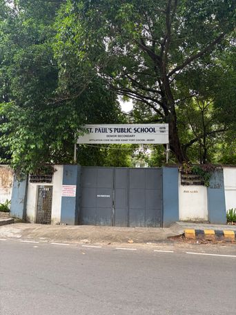 A large metal gate stands closed at the entrance to a property, flanked by white and blue walls under a canopy of leafy trees. A white sign is mounted above the gate, and a smaller notice is posted on the wall to the left. A yellow and black striped curb marks the edge of the road in front of the gate.