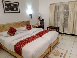 Room with two wooden beds with white bedding and red-patterned runners, a slatted wooden headboard, cream curtains, and a chrome coat stand.