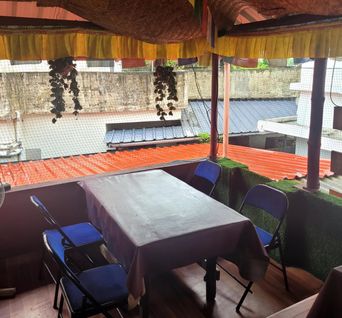 Rooftop dining table with folding blue chairs under a bamboo-thatch canopy, tin rooftops beyond.