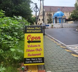 Yellow roadside sign for Tibetan Chef’s Restaurant, near Santa Cruz Basilica’s blue-domed shrine.
