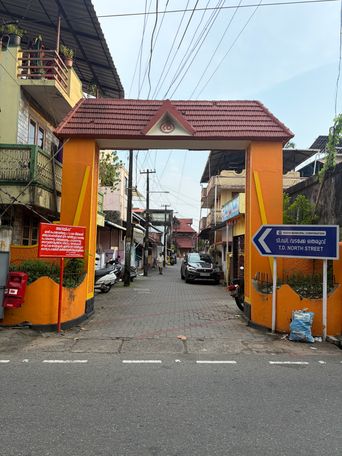 A wide paved street passes beneath a large orange gateway with a red tiled roof, leading into a narrow lane lined with buildings on both sides. A red sign stands on the left pillar of the gateway, and a blue directional sign is mounted on the right pillar. A black car drives toward the camera down the lane, while several scooters are parked along the left side. Overhead, numerous electrical wires crisscross the sky above the street.