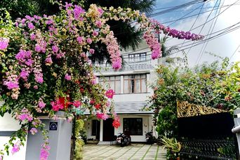 Two-storey white house with a tiled forecourt, its front wall and gateway covered in pink and magenta bougainvillea flowers.