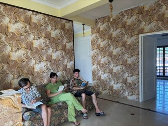 Sitting room with embossed brown-and-gold wallpaper on two walls, a patterned sofa where three people sit reading, and a glossy tiled floor leading to an open doorway.