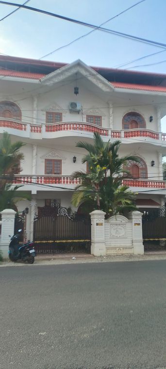A three-storey white building with red-tiled balconies and arched windows, behind a black iron gate and a palm tree, with a parked scooter on the street.