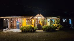 A single-storey house at night with a yellow facade, a carved wooden gable over the entrance, fairy lights strung along the eaves, and ferns and potted plants on a gravel forecourt.
