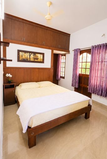 A bedroom with a double bed under a white cover, a wooden cabinet wall above it, purple curtains at an arched window, and a wall-mounted ceiling fan.