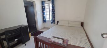 A small room with a double bed under a patterned white cover, blue curtains at a barred window, a dark wooden desk and chair on the left, and an open doorway to a bathroom.