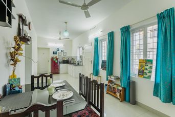 Open dining area with a glass-top table and dark wooden chairs, teal curtains at the windows, and a small kitchen with a red fridge beyond.