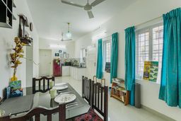 Open dining area with a glass-top table and dark wooden chairs, teal curtains at the windows, and a small kitchen with a red fridge beyond.