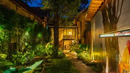 Courtyard garden lit at dusk between old tiled-roof buildings, with tables under a tree, a stone path and a rooster mural on one wall.