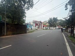 A paved road stretches into the distance under an overcast sky, with overhead power lines crisscrossing above. On the left, a high wall runs alongside dense trees, with a gate and a signboard visible beyond it. On the right, a person rides a motorcycle along the roadside, with more vehicles parked further down near buildings and trees. White lane markings divide the asphalt, and a few scattered leaves lie on the surface.