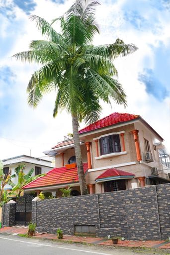 Two-storey house with red tiled roofs and orange columns, behind a dark brick-patterned boundary wall, with a tall palm tree growing beside it.