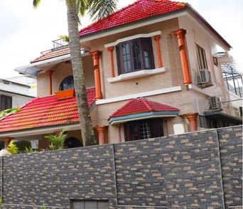 Two-storey house with brick-textured walls, orange columns and red tiled roofs, seen from the street past a tall palm tree and a dark stone-patterned boundary wall.