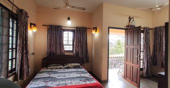 Bedroom with peach walls, a double bed with grey patterned bedding, patterned curtains, wall lamps and a ceiling fan, with an open wooden door onto a balcony.