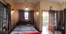 Bedroom with peach walls, a double bed with grey patterned bedding, patterned curtains, wall lamps and a ceiling fan, with an open wooden door onto a balcony.