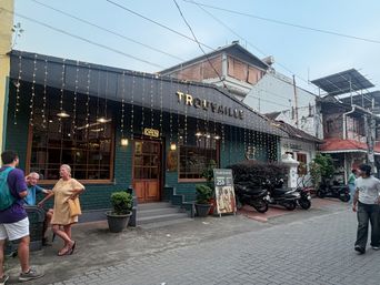 A street-level view of a green brick building with a dark awning and string lights hanging down, where several people are standing outside near the entrance and others walk along the paved sidewalk, with parked motorcycles and neighboring buildings visible in the background.