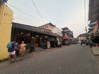 A street scene shows a row of buildings along a paved road. On the left, a dark green building with string lights across its front has people standing outside, including one person with a backpack and another in a light-colored dress. A signboard stands on the sidewalk near the entrance. Further down the street, other buildings line both sides, with parked motorcycles and a few people walking in the distance. Overhead wires stretch across the sky above the street.