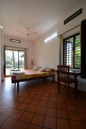 A room with a single bed under a ceiling fan, terracotta floor tiles, a wooden desk and chair by a window with iron grilles, and a glass door opening onto a green garden.