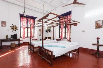 Room with two wooden four-poster beds with white bedding, red-patterned curtains at tall windows, and a red tiled floor.