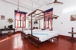 Room with two wooden four-poster beds with white bedding, red-patterned curtains at tall windows, and a red tiled floor.