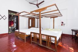 A white-walled room with red tiled floors, two wooden four-poster beds with white linen, a wooden wardrobe behind them, and a doorway on the left onto a veranda with a chair.