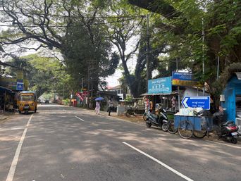 A paved road stretches through a tree-lined area with large trees forming a canopy overhead. On the left side of the road, a yellow auto-rickshaw is parked near the edge. Further down, two people walk along the roadside, one holding an umbrella. On the right side, several small roadside stalls and shops are set up under the trees, with various goods displayed. A blue cart with wheels is parked near one of the stalls, and a scooter is parked beside it. A blue directional sign with white text and an arrow stands near the carts. Overhead, numerous electrical wires crisscross above the road. The road surface has white lane markings, and shadows from the trees fall across the pavement.