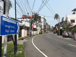 A blue directional sign with white text and an arrow points to the right along a curved asphalt road. The sign is mounted on a pole beside a grassy verge. Overhead, numerous electrical wires crisscross above the street. On both sides of the road are buildings, some with balconies and walls, and several palm trees are visible. Red flags hang from poles along the roadside. A scooter is parked on the right side near a wall, and a few motorcycles are further down the road. The sky is pale blue and clear.