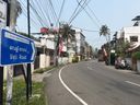A blue directional sign with white text and an arrow points to the right along a curved asphalt road. The sign is mounted on a pole beside a grassy verge. Overhead, numerous electrical wires crisscross above the street. On both sides of the road are buildings, some with balconies and walls, and several palm trees are visible. Red flags hang from poles along the roadside. A scooter is parked on the right side near a wall, and a few motorcycles are further down the road. The sky is pale blue and clear.