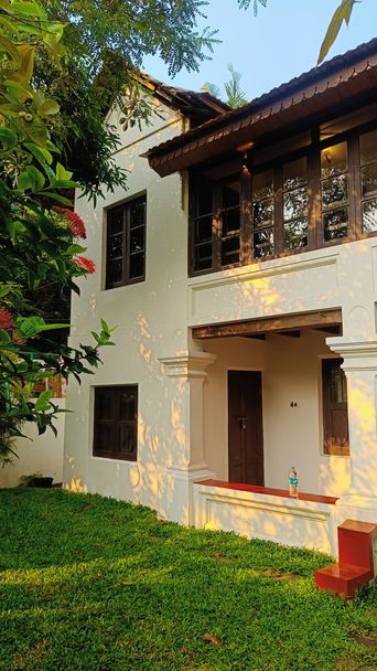 A white two-storey bungalow with dark-framed windows and a timber-roofed upper veranda, set behind lawn and a red-flowering shrub on the left.