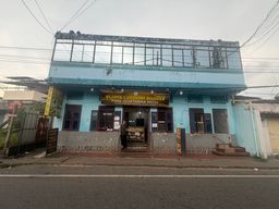 A two-story light blue building stands beside a paved road under an overcast sky, with overhead electrical wires crisscrossing above. The upper floor features a long row of windows behind a weathered metal railing, while the ground floor has several windows with dark frames and grilles. A person stands in the open doorway at the center, beneath a large horizontal sign mounted above the entrance. Additional vertical signs are positioned on either side of the building, and a short flight of steps leads up to the entrance from the sidewalk.