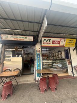 A street-level view of two adjacent sweet shops under a shared corrugated roof. On the left, a shop entrance features a glass display counter on wheels, with a person seated inside near a water dispenser. A wooden table and two red plastic stools sit outside. On the right, another shop has shelves stocked with packaged goods and a refrigerated display case filled with items. A vertical sign stands between the two entrances, and several signs are mounted above the storefronts.