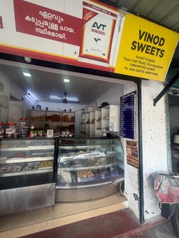 The entrance to a sweet shop with a glass display counter filled with assorted sweets and snacks. Shelves behind the counter hold more packaged goods and jars. A yellow sign above the entrance displays the shop name and contact details, while a red sign with text in a regional script is visible on the left. A menu board stands on the wall to the right, and a poster is attached to the pillar beside the entrance.