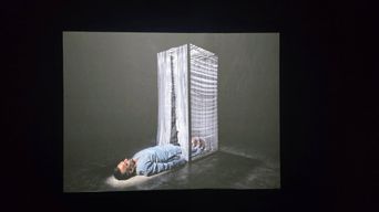 A projected image in a dark room: a person lying on their back on the floor beside a tall glass case with a curtain hanging inside it.