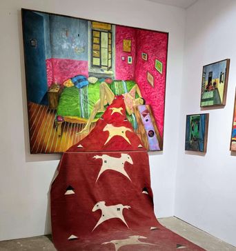 A gallery wall hung with a large painting of a bedroom in hot pink, green and blue, with a dark red rug patterned with white deer spilling down the wall onto the floor in front of it.