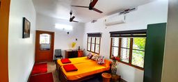 Bedroom with an orange bedspread on a double bed and red cushions, an air conditioner on the wall, a ceiling fan, two barred windows and red rugs on the tiled floor.