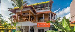 Two-storey white building with an orange-tiled roof, a wooden balcony on the upper floor and an orange sign below it, set among palm trees under a blue sky.